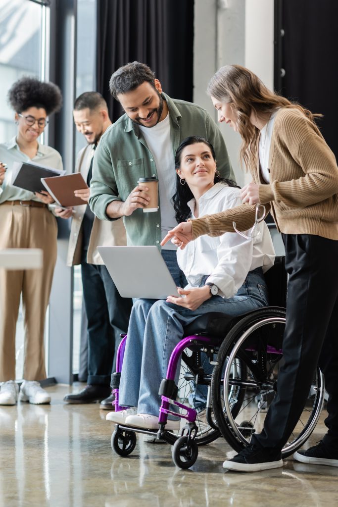 disabled woman in wheelchair discussing startup project with happy interracial team in coworking